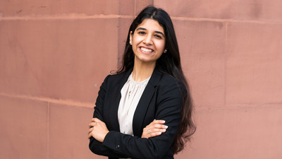 Eine lächelnde Person trägt eine altrosa farbene Bluse sowie einen schwarzen Blazer und steht vor einer Steinwand. Die Person heißt Tushna Vevaina.