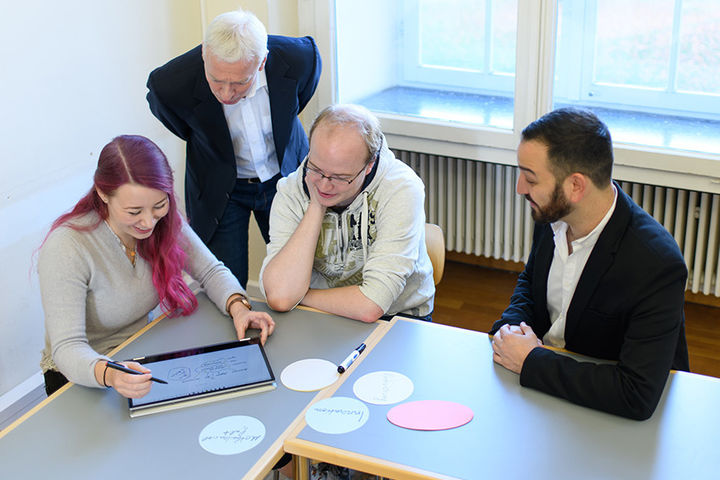 Eine junge Frau mit pinken Haaren zeigt drei interessierten Männern eine Skizze auf ihrem Laptop.