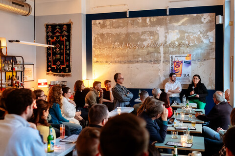 Viele Personen sitzen in einer Bar und hören einer vortragenden Person zu.