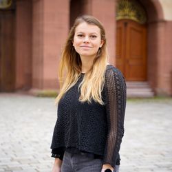 Eine junge Studentin steht vor dem Mannheimer Schloss. Sie trägt lange, blonde Haare und ein schwarzes Oberteil.