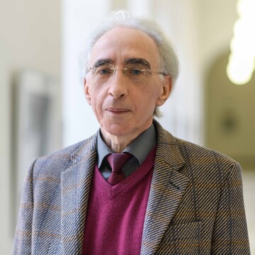 Prof. Dr. Christoph Weiß trägt eine braunen Anzug mit einem violetten Pullunder. Er hat kurze graue Haare und trägt eien rahmenlose Brille.