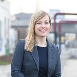 Portrait von Julia Derkau. Sie trägt ein schwarzes Oberteil sowie einen grauen Blazer und steht vor einem Gebäude.
