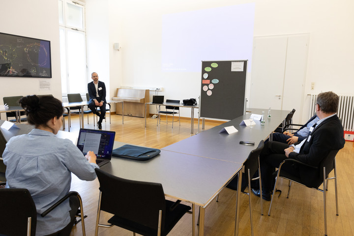 Großer Seminarraum mit Tischen, die im Viereck stehen. In einer Ecke sitzt Prof. Dr. Heiner Stuckenschmidt, vorne im Bild sieht man zwei Teilnehmende des Workshops. Im Hintergrund steht eine Pinnwand mit kleinen Karten darauf. 