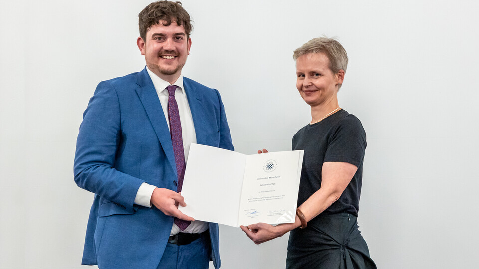 Dr. Nico Halkenhaeuser, blaues Jacket und dunkle, kurze Haare, erhält die Urkunde für den Lehrpreis von Prof. Dr. Cornelia Ruhe, die ein schwarzes Kleid trägt. Beide stehen nebeneinander und lächeln in die Kamera.
