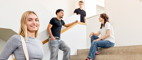 Eine Studentin mit blonden Haaren und einer Jute-Tasche steht auf der Treppe in B6. Im Hintergrund unterhalten sich 3 Studierende.
