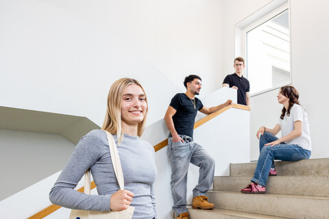 Eine Studentin mit blonden Haaren und einer Jute-Tasche steht auf der Treppe in B6. Im Hintergrund unterhalten sich 3 Studierende.