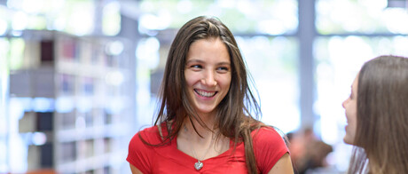 Zwei weiblich gelesene Studierende stehen an einem Bibliotheksregal und lachen sich an. Die Person, die von vorne zu sehen ist, trägt ein rotes Oberteil. Die andere Person steht im Profil, hat ein dunkelrotes T-Shirt und hält Ordner in ihrem Arm. Beide haben lange braune Haare.