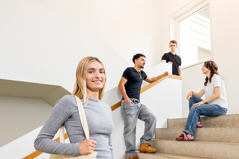 Eine Studentin mit blonden Haaren und einer Jute-Tasche steht auf der Treppe in B6. Im Hintergrund unterhalten sich 3 Studierende.