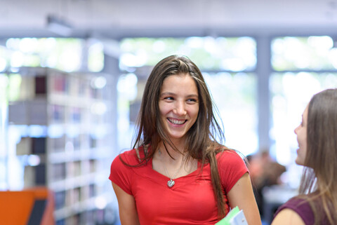 Zwei weiblich gelesene Studierende stehen an einem Bibliotheksregal und lachen sich an. Die Person, die von vorne zu sehen ist, trägt ein rotes Oberteil. Die andere Person steht im Profil, hat ein dunkelrotes T-Shirt und hält Ordner in ihrem Arm. Beide haben lange braune Haare.