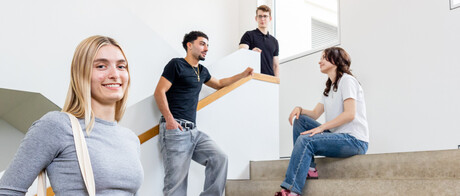 Eine Studentin mit blonden Haaren und einer Jute-Tasche steht auf der Treppe in B6. Im Hintergrund unterhalten sich 3 Studierende.