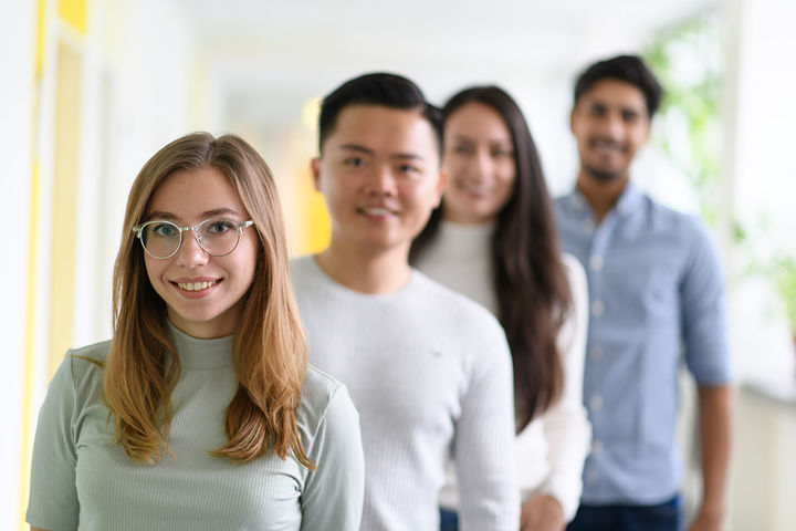 Zwei Studentinnen und zwei Studenten stehen schräg hintereinander. Nur die vordere Studentin ist scharfgestellt. Sie trägt lange blonde Haare, eine silberne Brille und einen mintfarbenen Rollkragenpullover.