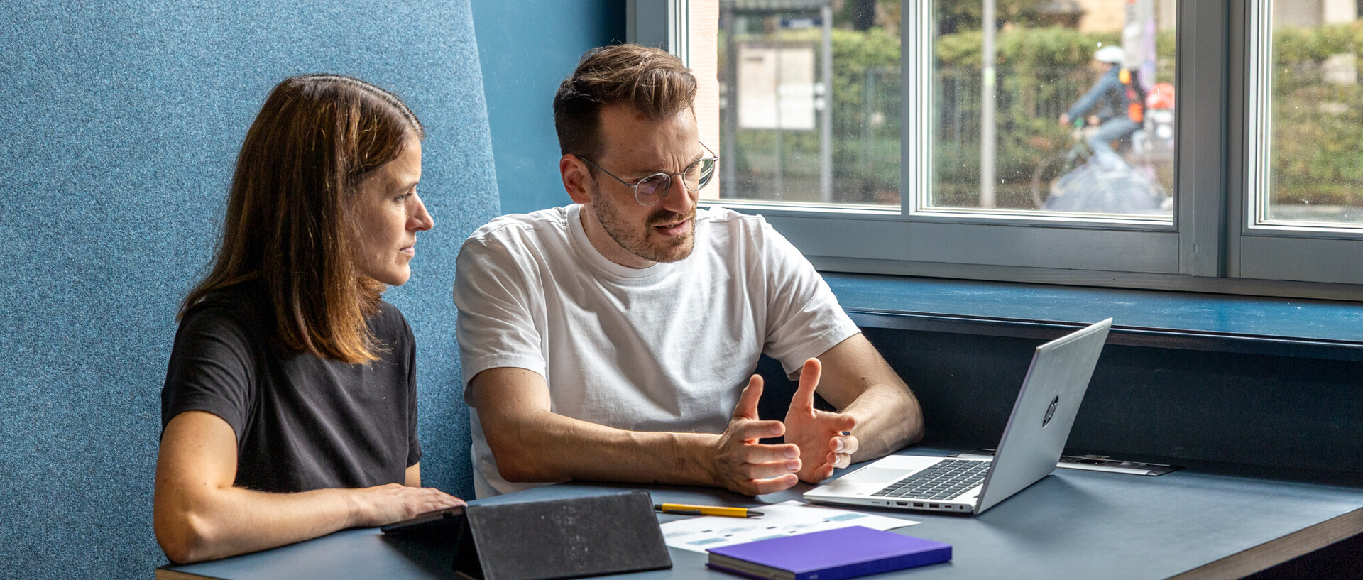 A man and a woman are sitting at a table together, looking intently at a laptop.