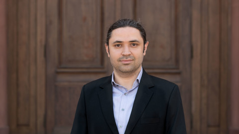 Mohammad Zenouzzadeh. He is wearing a black suit and is is standing in front of a brown door.