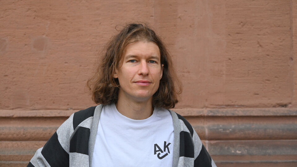 Richard Rohrmoser steht vor einer rötlich-braunen Wand. Er trägt ein weißes Shirt, darüber eine grau-Schwarz gestreifte Stoffjacke. Er hat schulterlanges braun-blondes Haar. 