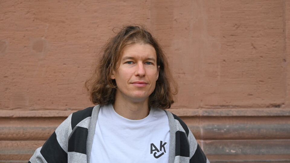 Richard Rohrmoser steht vor einer rötlich-braunen Wand. Er trägt ein weißes Shirt, darüber eine grau-Schwarz gestreifte Stoffjacke. Er hat schulterlanges braun-blondes Haar.