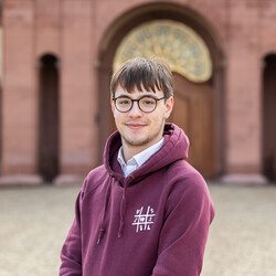 Franz Stengel, mit kurzen schwarzen Haaren und Brille, trägt einen weinroten Pullover mit dem Logo der Fachschaft und lächelt freundlich in die Kamera.