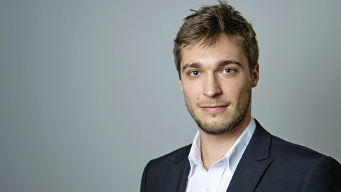 Carlo Birkholz. He is wearing a black suit and is standing in front of grey wall.