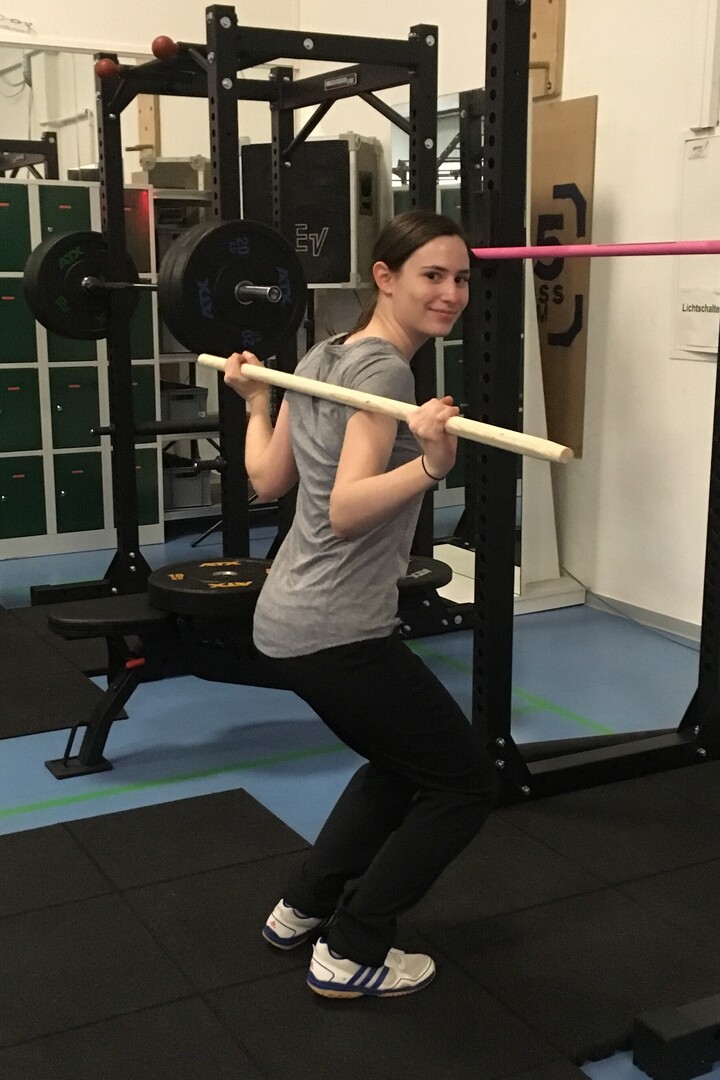 Eine junge Frau trainiert mit einer Holzstangen ihre Rückenpartie im Fitnessstudio