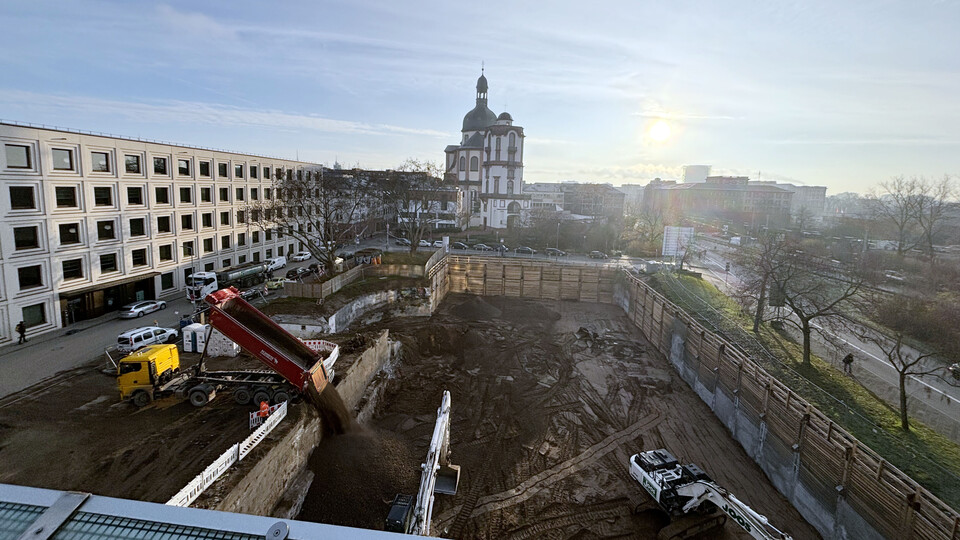 Im Vordergrund die Baustelle in A5 mit großer Baugrube, im Hintergrund die Jesuitenkirche Mannheim bei Sonnenaufgang. Ein Lkw kippt Erde in die Grube, daneben zwei Bagger. Links das modernes Gebäude B6, rechts Bäume und eine Straße.