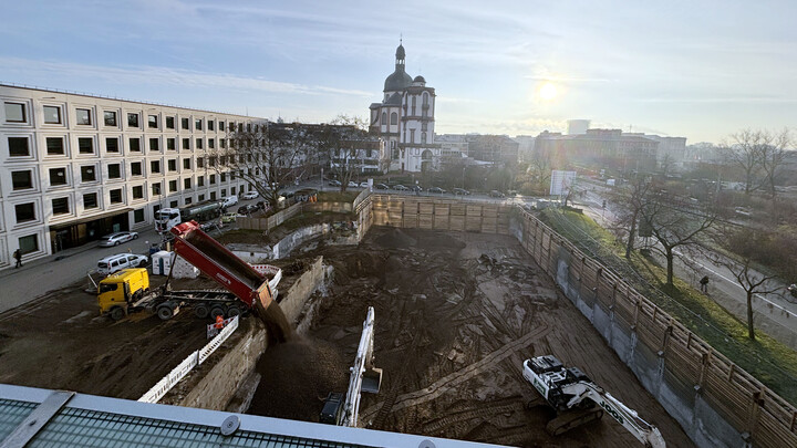 Im Vordergrund die Baustelle in A5 mit großer Baugrube, im Hintergrund die Jesuitenkirche Mannheim bei Sonnenaufgang. Ein Lkw kippt Erde in die Grube, daneben zwei Bagger. Links das modernes Gebäude B6, rechts Bäume und eine Straße.
