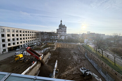 Im Vordergrund die Baustelle in A5 mit großer Baugrube, im Hintergrund die Jesuitenkirche Mannheim bei Sonnenaufgang. Ein Lkw kippt Erde in die Grube, daneben zwei Bagger. Links das modernes Gebäude B6, rechts Bäume und eine Straße.
