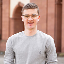 A portrait of Sören Hengstberger. He is standing in the Ehrenhof, smiling, wearing glasses and a gray sweater, and has short blond hair.