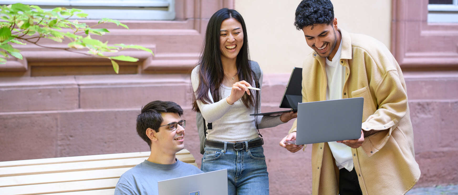 Drei Studierende halten Laptops in ihren Händen und lachen.