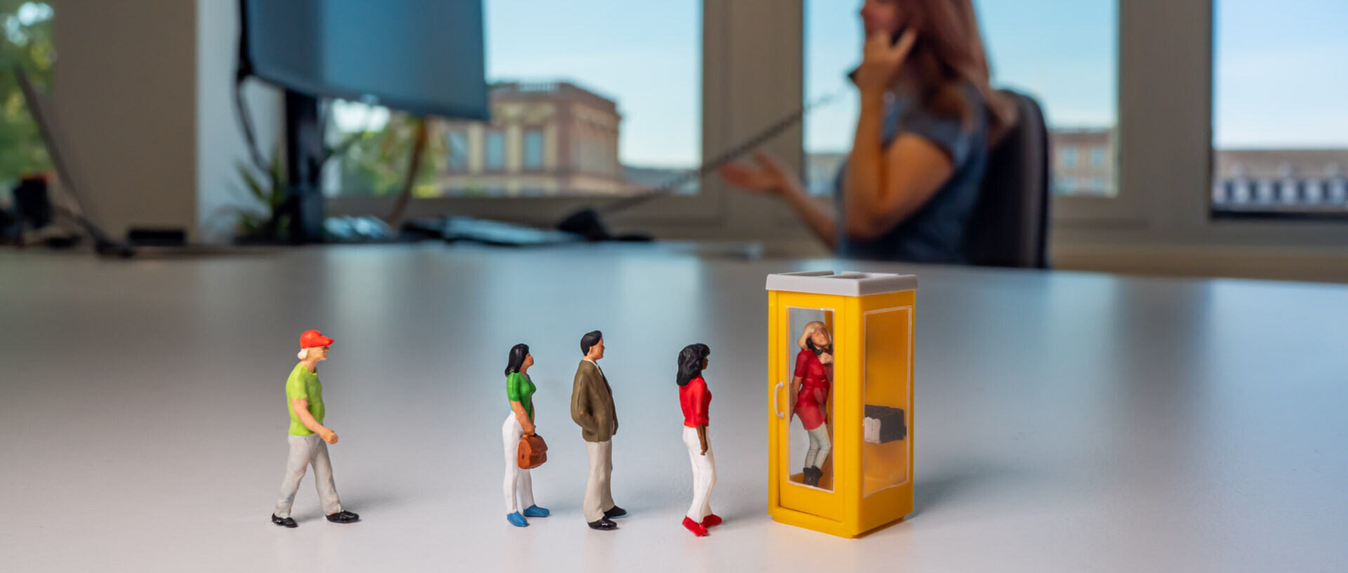 Das Foto zeigt im Hintergrund eine leicht verschwommene Person am Telefon, im Vordergrund telefoniert eine Miniaturfigur in einer Telefonzelle, weitere Miniaturfiguren stehen Schlange.