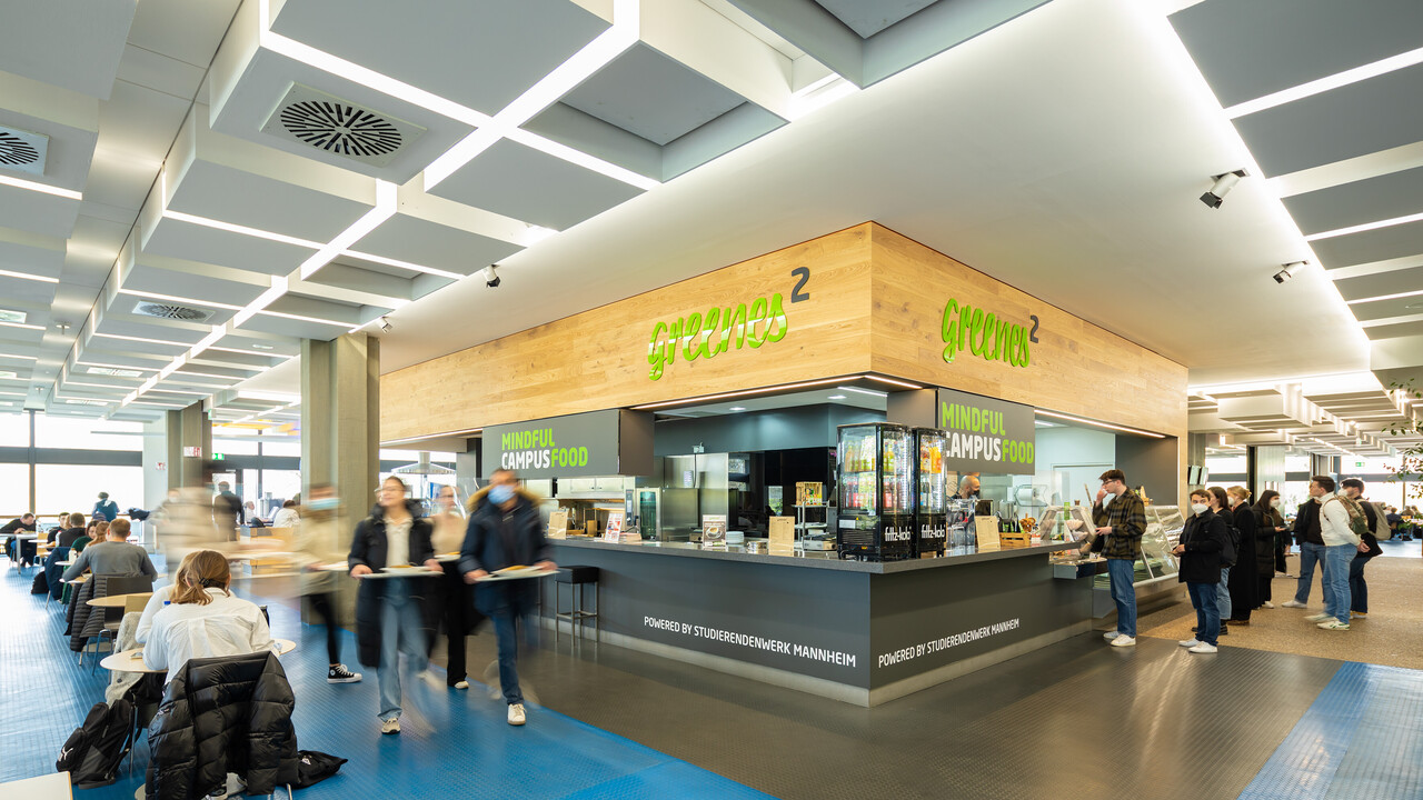 The greenes² at the Mensa Eine moderne Theke aus Holz in der Mensa. Auf der Theke steht mit greller, grüner Schrift "greenes²" und "mindful campus food".