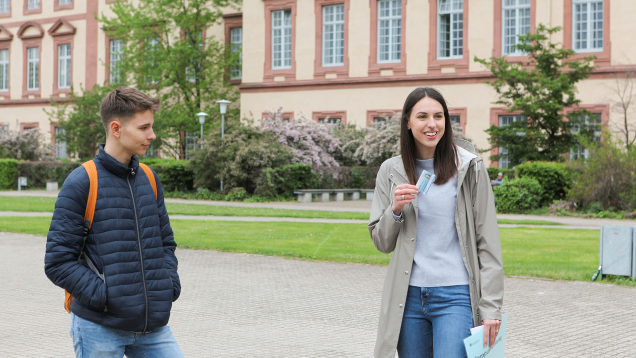 Eine junge Frau macht eine Campus-Führung. Sie steht auf der Mensawiese und hält einen Studierendenausweis hoch. Neben ihr ein Student.