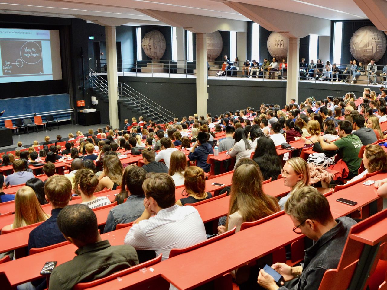 Studierende sitzen in einem roten Hörsaal. Die Studierenden hören einem Vortrag zu.