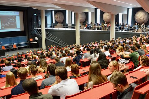 Studierende sitzen in einem roten Hörsaal. Die Studierenden hören einem Vortrag zu.