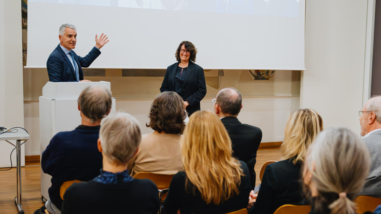 Das Bild zeigt den Rektor der Universität in einem blauen Anzug am Rednerpult. Neben ihm steht die schwarz gekleidete Festrednerin. Beide lachen. Im Vordergrund des Bildes sieht man Hinterköpfe einiger Zuhörenden.