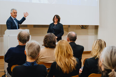 Das Bild zeigt den Rektor der Universität in einem blauen Anzug am Rednerpult. Neben ihm steht die schwarz gekleidete Festrednerin. Beide lachen. Im Vordergrund des Bildes sieht man Hinterköpfe einiger Zuhörenden.