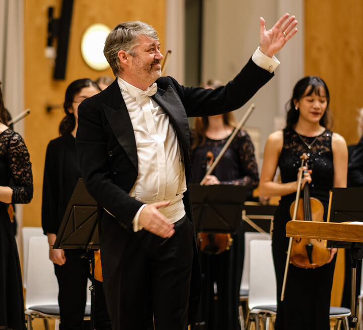 Auftritt des Uni Orchesters. Dirigent steht vorne und hinter ihm befinden sich Studierende mit Instrumenten