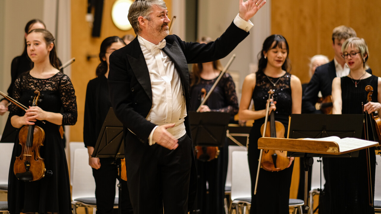 Auftritt des Uni Orchesters. Dirigent steht vorne und hinter ihm befinden sich Studierende mit Instrumenten