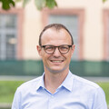 Eine lächelnde Person trägt ein blau-weiß gemustertes Hemd sowie eine beige-farbene Hose mit einem braunen Gürtel und steht auf der Mensawiese vor dem Schloss. Die Person heißt Moritz Kuhn.