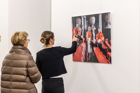 Zwei weiblich gelesene Personen stehen vor einem Bild der Spitzensport-Vernissage. Eine davon zeigt auf ein Portrait. 