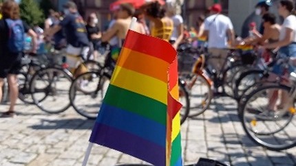 Fahrrad mit Regenbogen-Fahne im Vordergrund und im HIntergrund viele Menschen auf Fahrrädern.