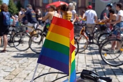 Fahrrad mit Regenbogen-Fahne im Vordergrund und im HIntergrund viele Menschen auf Fahrrädern.