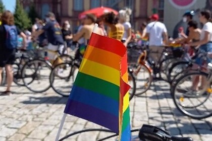 Fahrrad mit Regenbogen-Fahne im Vordergrund und im HIntergrund viele Menschen auf Fahrrädern.