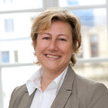 Portrait von Angelika Wöllstein. Sie trägt eine weiße Bluse und einen braunen Blazer und steht lächelnd vor einer Fensterfront.
