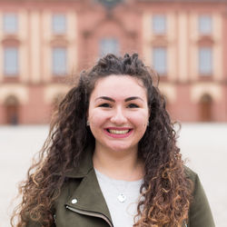 Cristina Solaro has curly long brown hair. She is wearing a light-colored top and an olive-colored jacket and is standing in front of the castle.