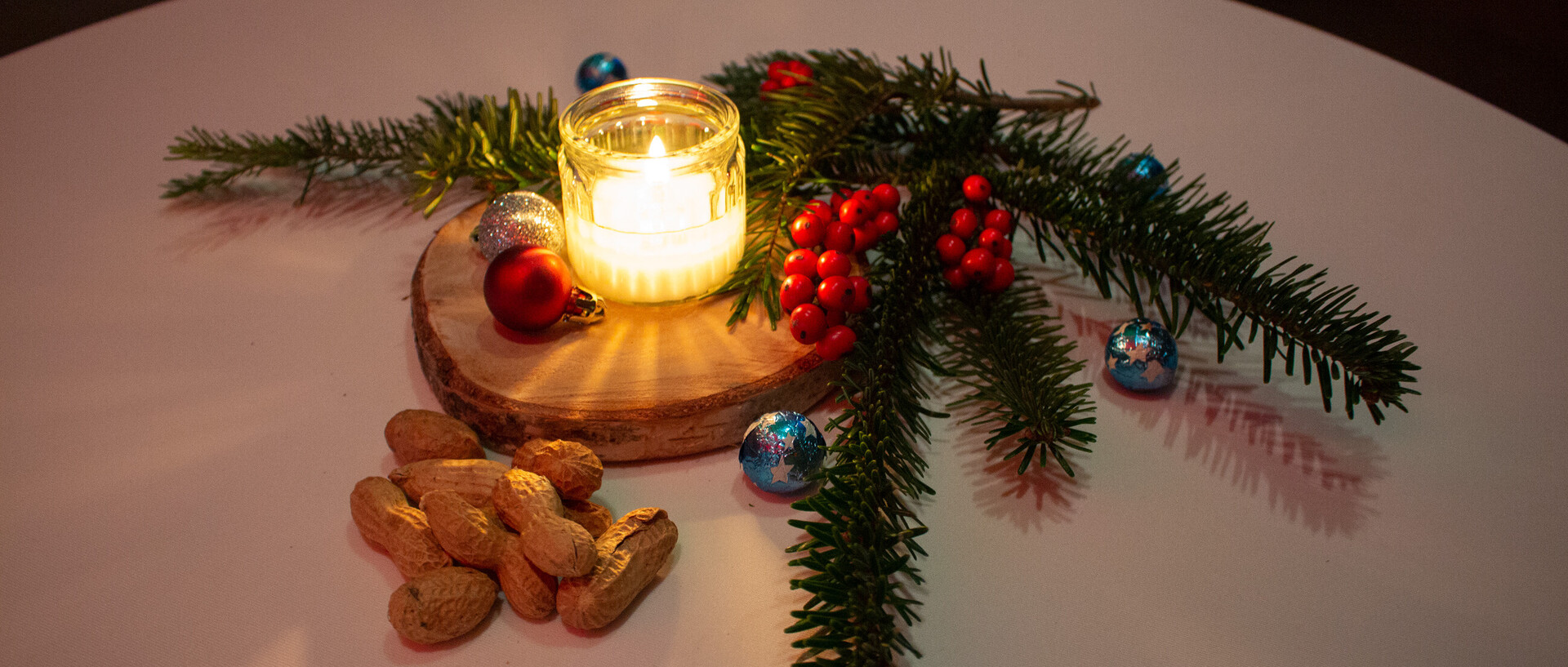 Auf einem Tisch scheint eine Weihnachtskerze. Darum ein Tannenzweig, Nüsse und kleine bunte Weihnachtskugeln.