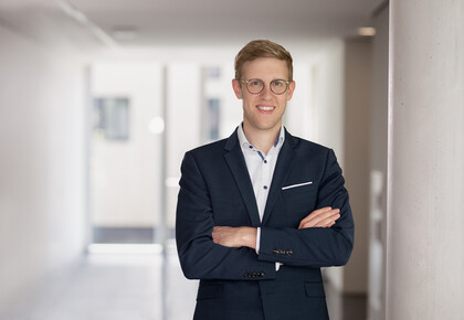 Jens Förderer. He is wearing a black suit and is standing inside a white walled building.