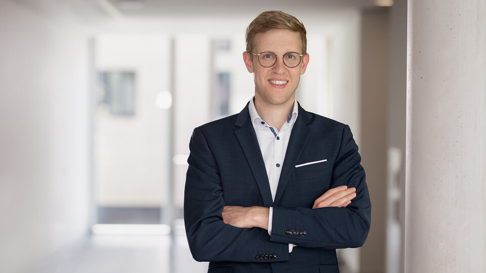 Jens Förderer. He is wearing a black suit and is standing inside a white walled building.