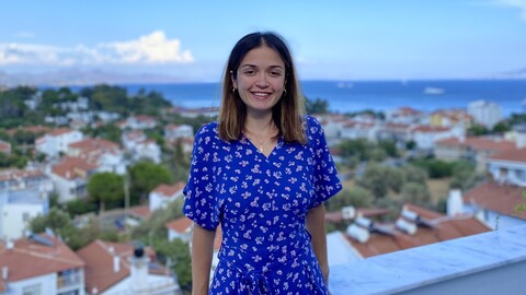Hatice Dedetaș Şatır. She is wearing a blue dress and standing in front of and above houses and the sea.