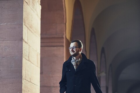 Mann mit Brille, einer dunkelblauen Stoffhose, einem schwarzen langem Mantel und einem gestreiften Schaal steht schwebend über dem Boden mit einem Koffer in der Hand in einem der Gänge vor der Universität.