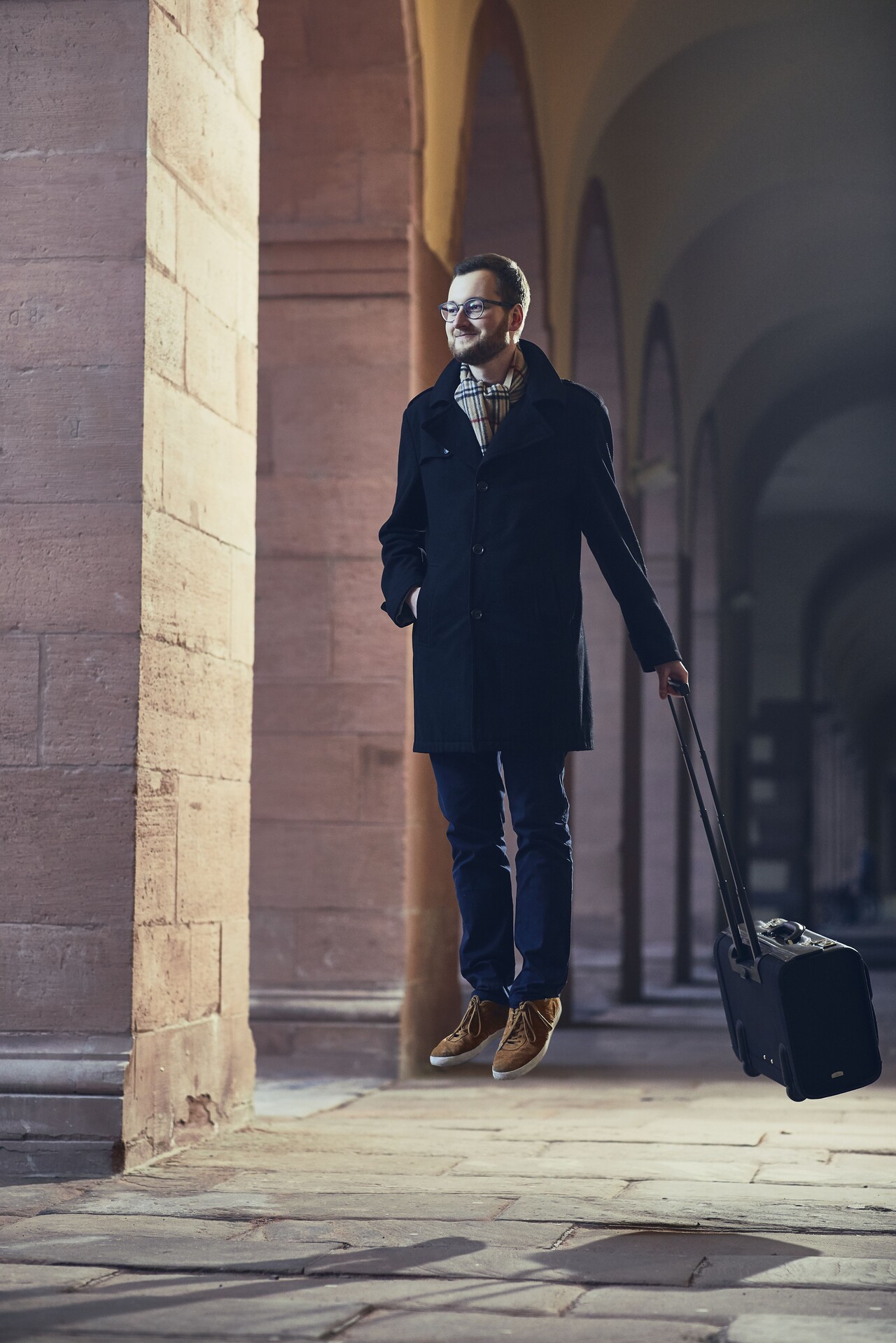 Mann mit Brille, einer dunkelblauen Stoffhose, einem schwarzen langem Mantel und einem gestreiften Schaal steht schwebend über dem Boden mit einem Koffer in der Hand in einem der Gänge vor der Universität.