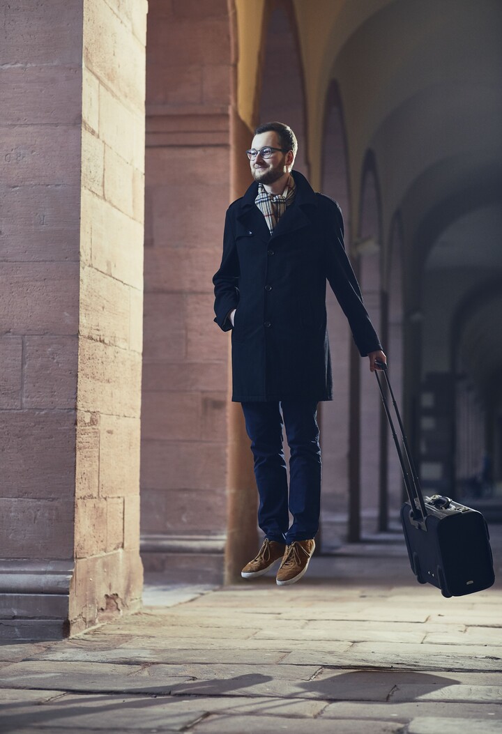 Mann mit Brille, einer dunkelblauen Stoffhose, einem schwarzen langem Mantel und einem gestreiften Schaal steht schwebend über dem Boden mit einem Koffer in der Hand in einem der Gänge vor der Universität.